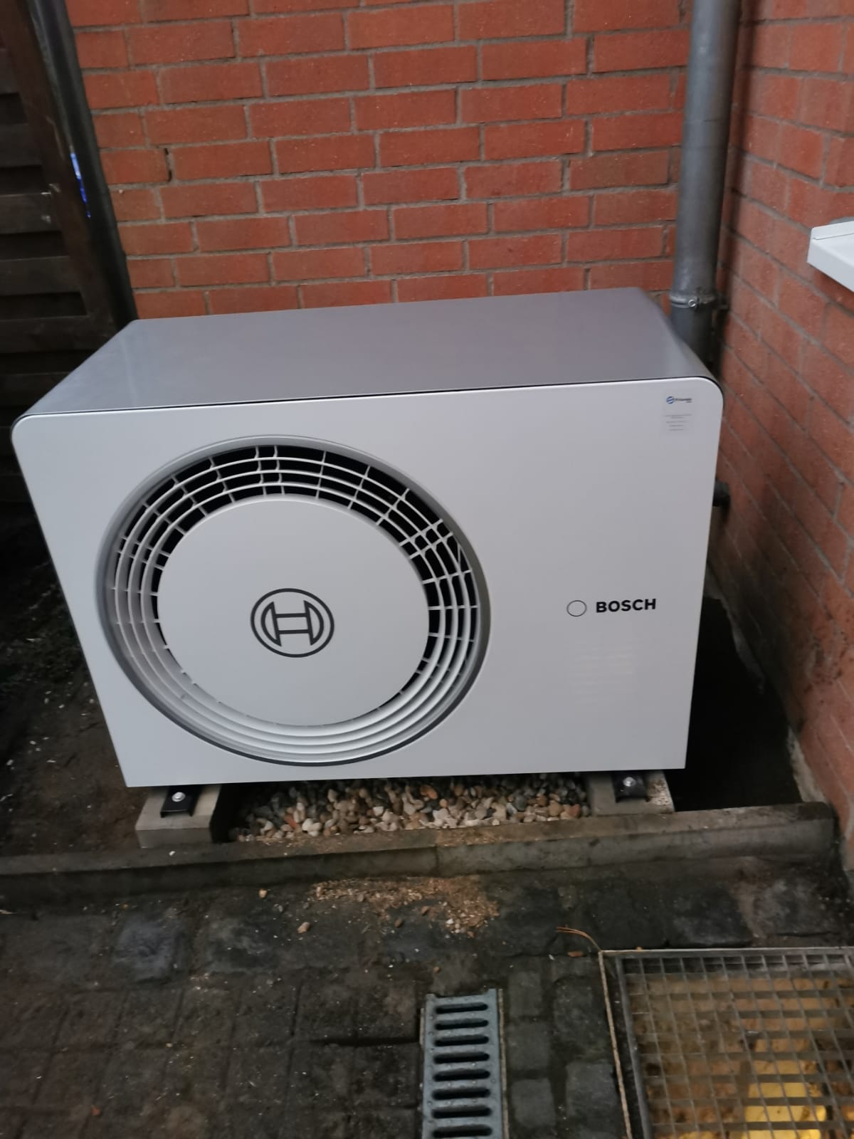 Bosch-Wärmepumpe auf Betonfundament mit Kiesbett vor einer roten Ziegelwand nahe einem Fenster und Wasserablauf.